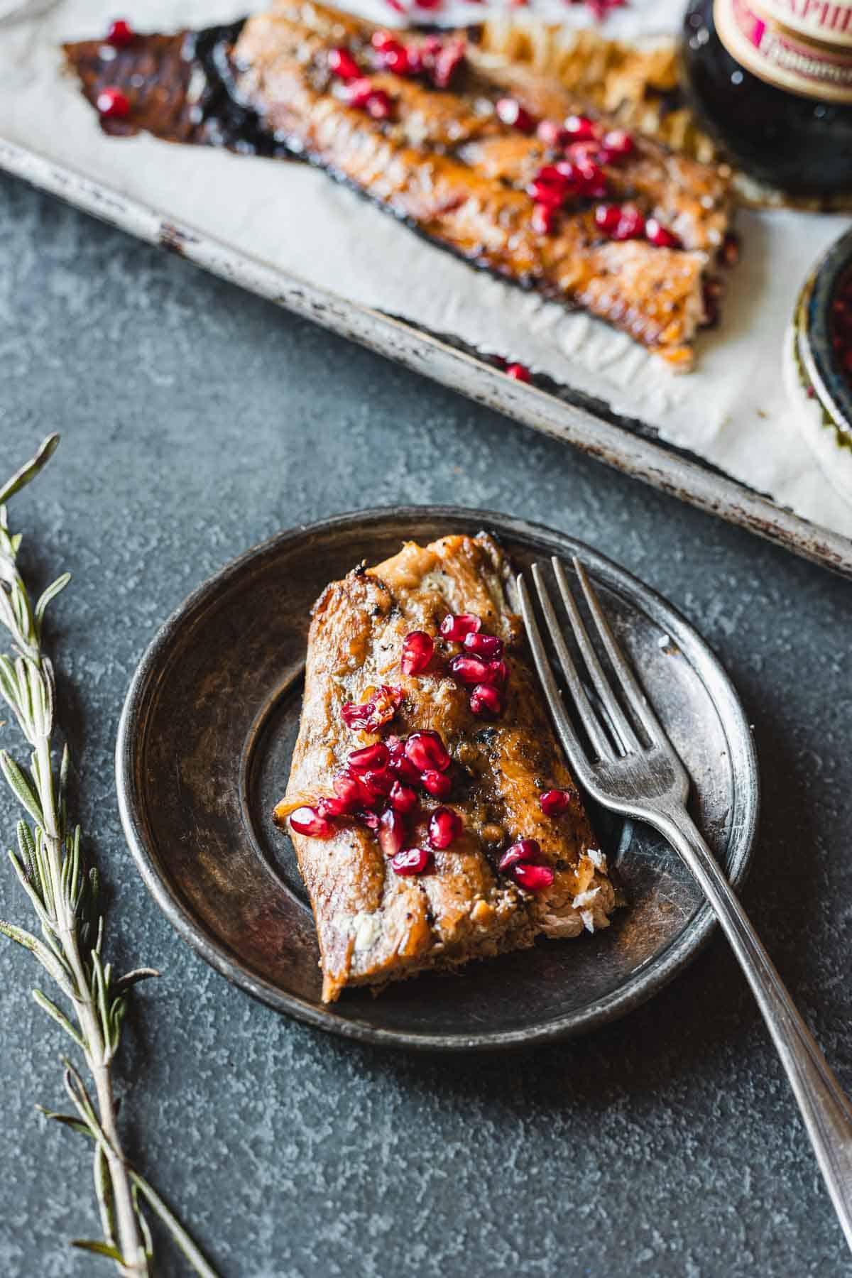 A plate with a piece of oven-baked salmon garnished with pomegranate seeds and a fork resting on the side. In the background, there is another piece of fish on a baking tray, and a sprig of rosemary is placed next to the plate.