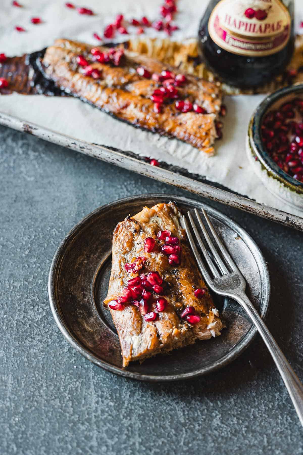 A serving of oven-baked salmon topped with pomegranate seeds is displayed on a round, gray plate with a fork. A larger portion of the glazed salmon and a dish with more pomegranate seeds can be seen in the background on a baking sheet. A bottled condiment is also visible.