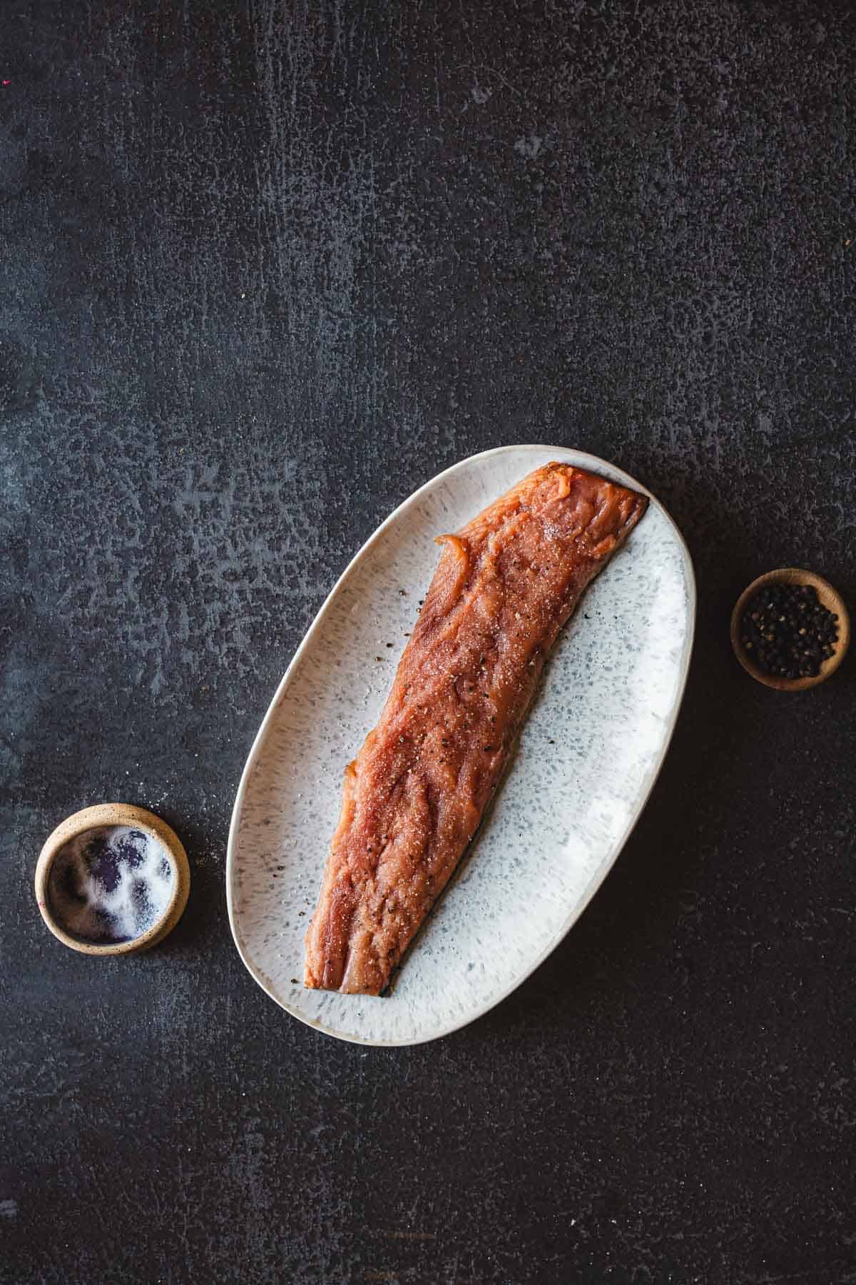 A grilled piece of pomegranate glazed salmon fillet is placed on an oval white plate. A small dish containing black peppercorns and a glass of dark beverage, possibly beer or coffee, are positioned beside the plate on a dark textured surface.