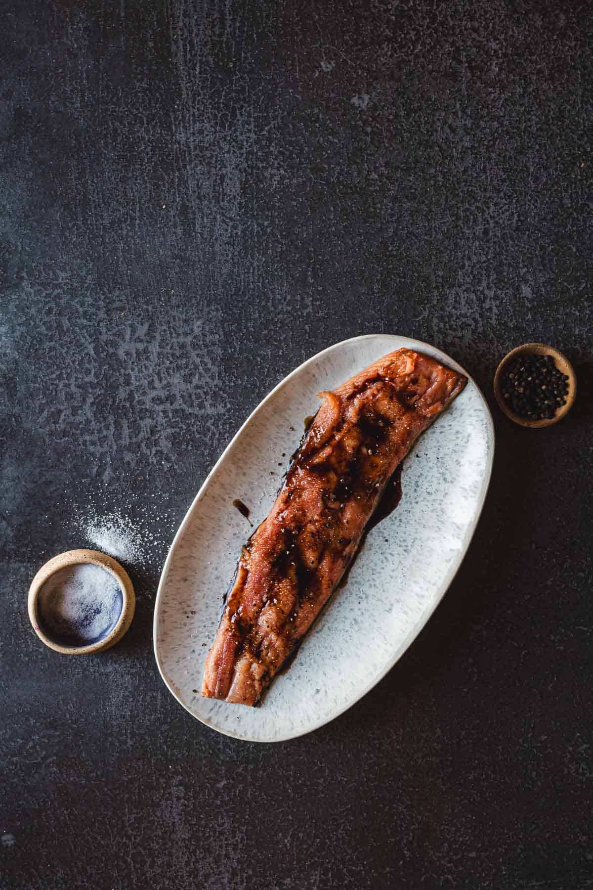 An oval plate holds a pomegranate-glazed salmon fillet on a dark surface. The fish has grill marks and is drizzled with the luscious glaze. A small round dish of salt and another of black peppercorns are placed nearby on the table.