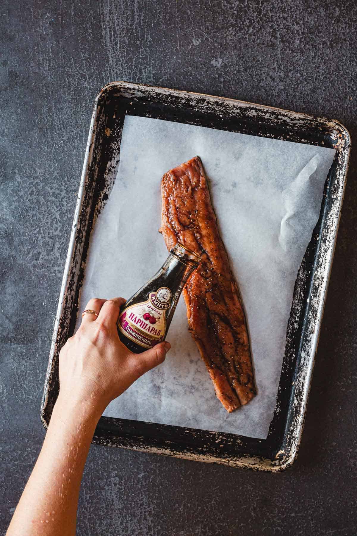 A hand is pouring a bottle of marinade over a raw salmon fillet on a parchment-lined baking tray, ready to become pomegranate glazed salmon. The background is a dark, textured surface.