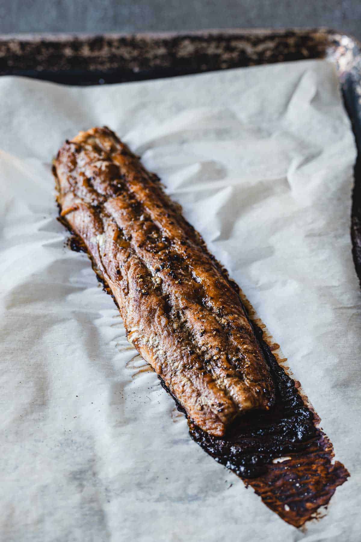 A cooked fillet of oven-baked salmon, seasoned and browned, rests on a sheet of parchment paper on a baking tray. The fish appears caramelized with darker, crispy edges. The parchment paper has absorbed some oils and juices, hinting at a rich flavor profile without overwhelming the senses.