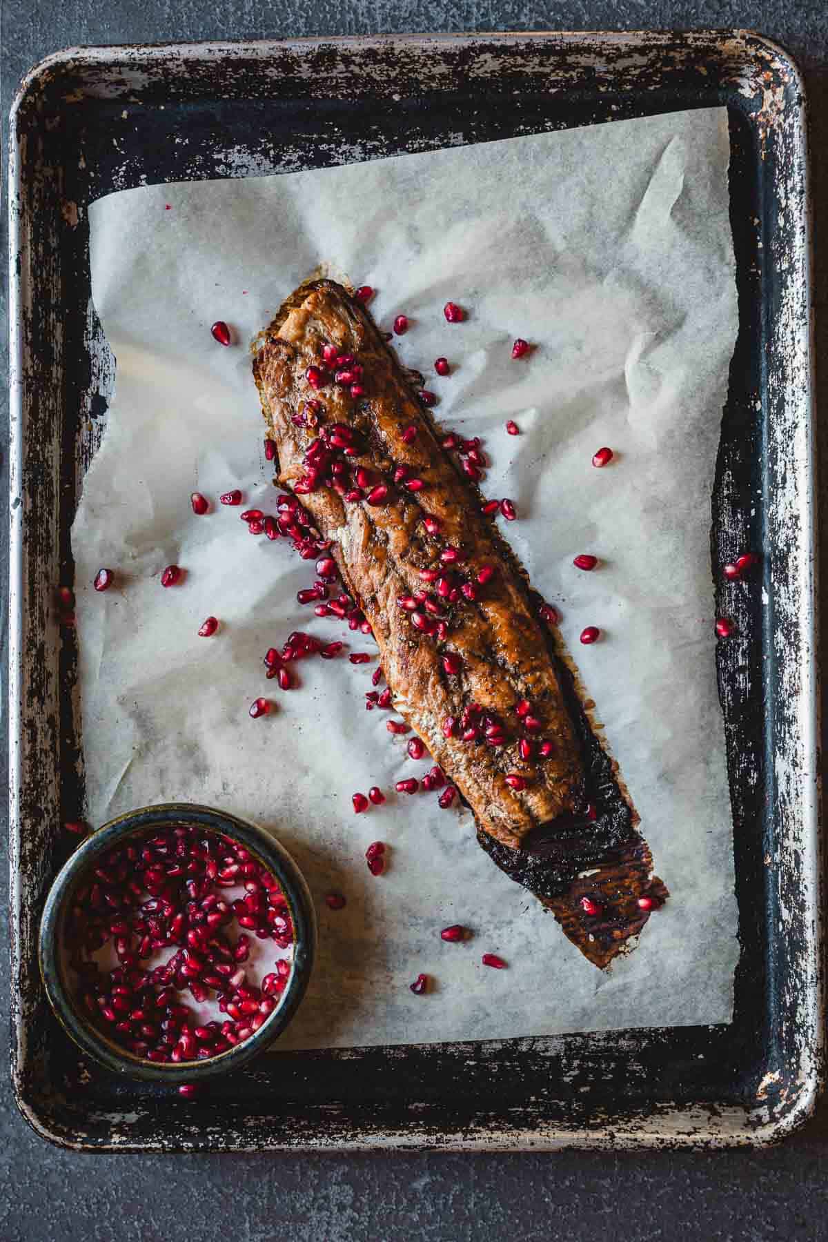 A roasted piece of pomegranate glazed salmon, topped with pomegranate seeds, is placed on parchment paper on a weathered baking sheet. A small bowl filled with additional pomegranate seeds sits in the bottom-left corner of the baking sheet.