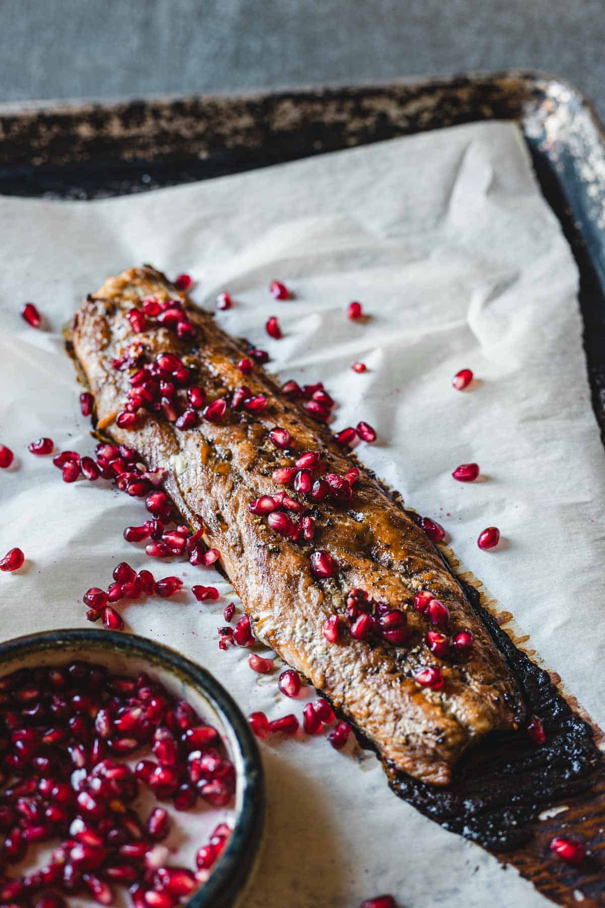 A cooked piece of salmon rests on a sheet of parchment paper placed on a baking tray. The fish is oven-baked and garnished with pomegranate seeds. A small bowl of pomegranate seeds is partially visible in the lower-left corner of the image.