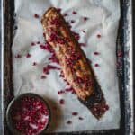 A roasted pork tenderloin, pomegranate glazed and topped with scattered seeds, rests on a sheet of parchment paper on a dark, rustic baking tray. A small bowl filled with more pomegranate seeds is situated to the left of the tenderloin.