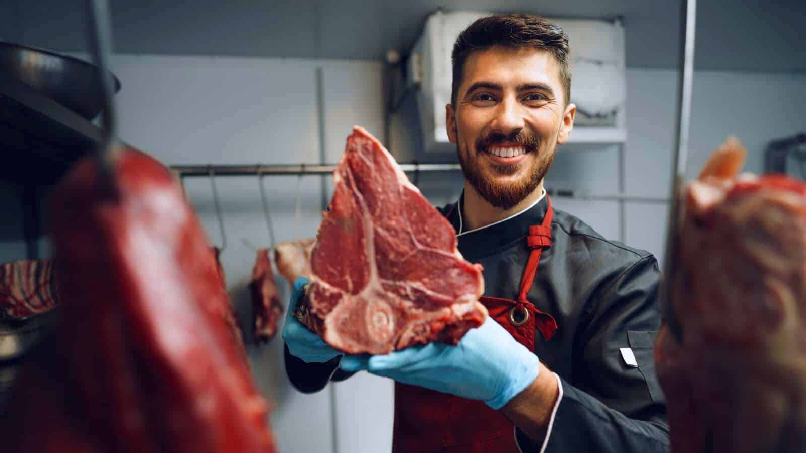 A man wearing a black jacket and blue gloves holds a large piece of raw meat, smiling. He is in a room with other pieces of meat hanging around him.