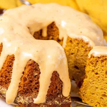 A round, golden-brown pumpkin bundt cake with a thick layer of creamy glaze drips on the top and sides. A slice has been cut and placed in front, showcasing the moist interior of the cake. Yellow fabric is partially visible in the background, creating an auto draft of cozy autumn vibes.