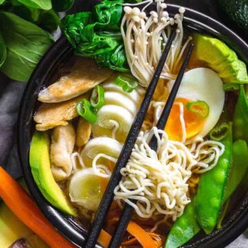 A bowl of ramen filled with noodles, a halved boiled egg, sliced green onions, snow peas, and enoki mushrooms. It also contains slices of chicken, carrot, avocado, and baby bok choy. This dish is surrounded by fresh vegetables and mushrooms on a gray cloth—an Asian recipe that's shockingly easy to make.
