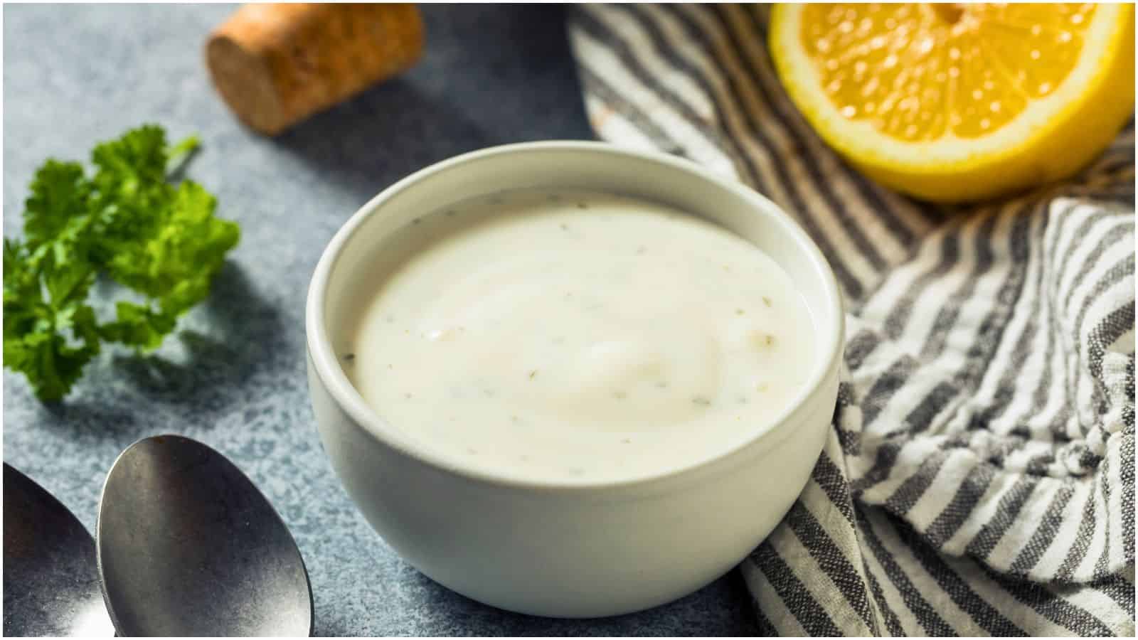 A bowl of creamy ranch dressing, a quintessential favorite among American foods, is placed on a table next to a striped cloth napkin, a halved lemon, a cork, and a sprig of parsley. Two spoons are also visible in the foreground.