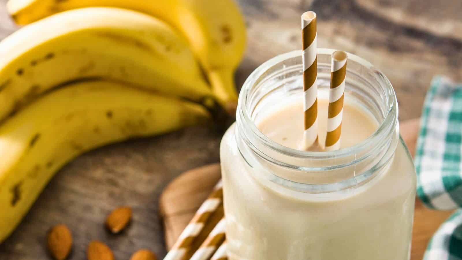 A close-up of a creamy smoothie in a glass jar with two striped paper straws. In the background, a bunch of ripe bananas is visible. A green and white checkered cloth is placed to the right, alongside a few scattered almonds. The surface is wooden.