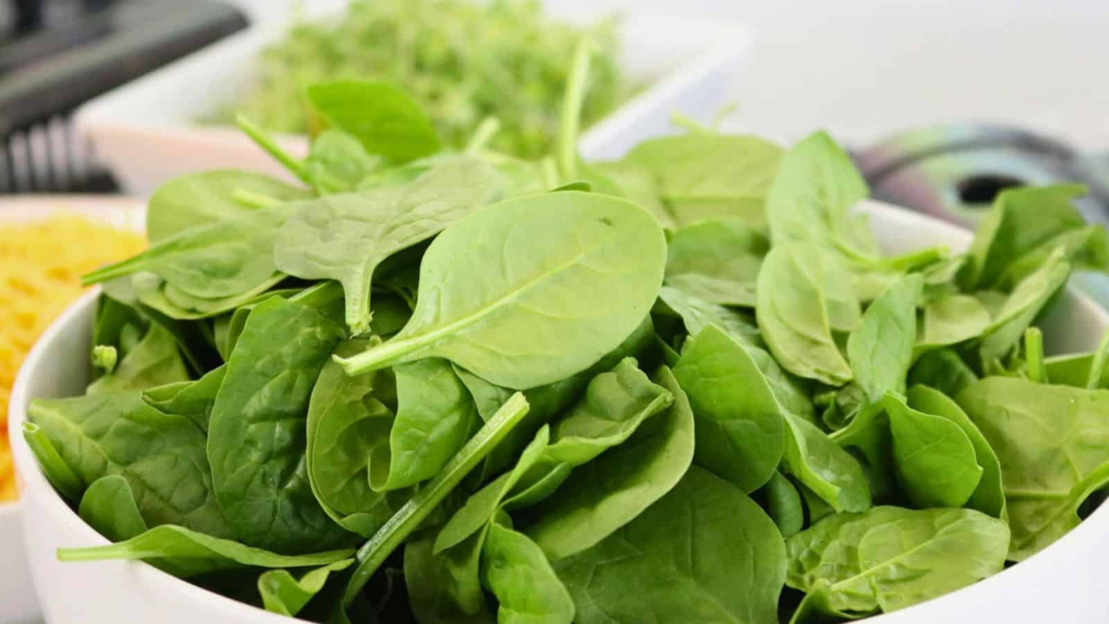 A white bowl filled with fresh green spinach leaves sits on a table. Other food items are blurred in the background, including some leafy greens and what appears to be grated cheese.