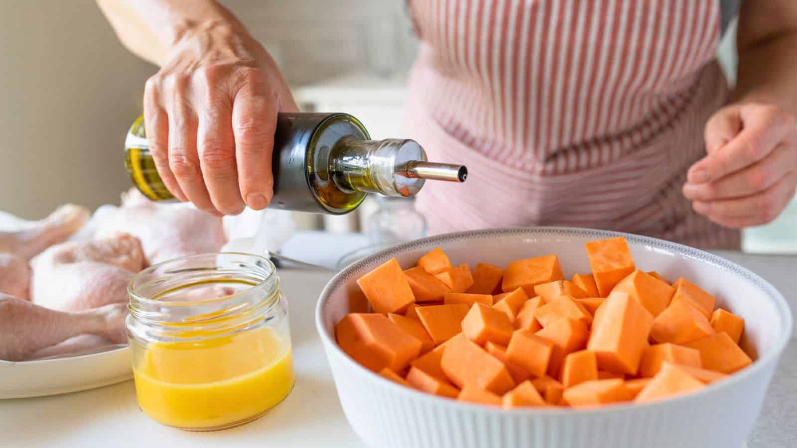 A person wearing a striped apron pours olive oil onto a bowl of cubed sweet potatoes. Nearby, an open jar with Auto Draft containing a yellow liquid, presumably melted butter, sits next to a plate with raw chicken drumsticks.