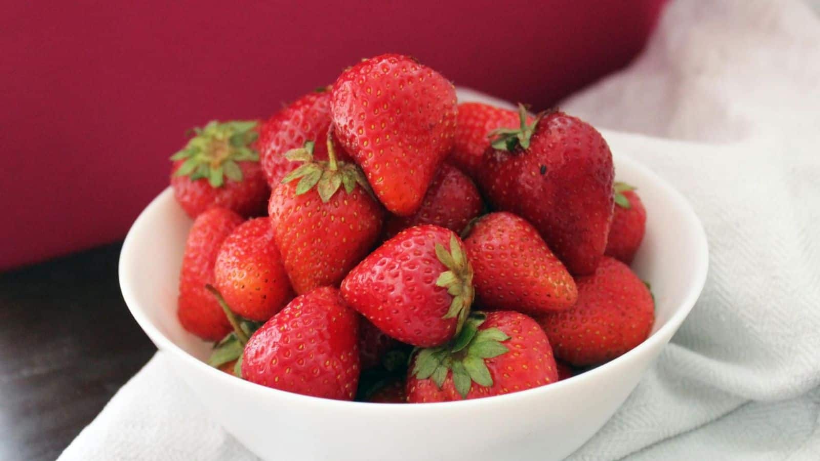 A white bowl filled with fresh strawberries is placed on a white fabric with a pink background. The strawberries are vivid red with green leaves, showing their fresh and ripe quality.