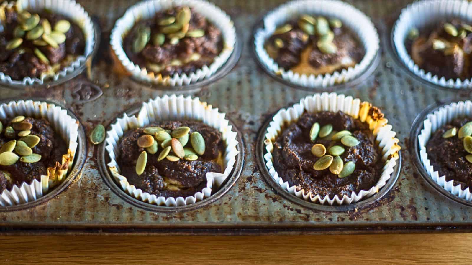 A muffin tin containing eight chocolate muffins lined with white paper cups, each topped with pumpkin seeds. The tin shows signs of use, with some residue and slight rust, and sits on a wooden surface, reminding you that you’re missing out on trying new recipes.