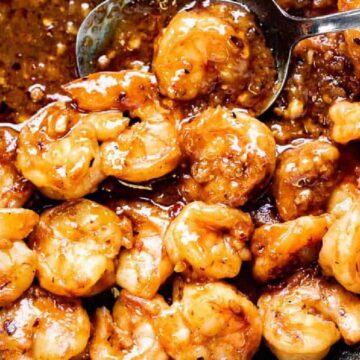 A close-up of cooked shrimp in a pan with a thick, dark brown sauce. The shrimp are evenly coated, and a spoon is seen lifting some sauce from the pan. This savory seafood preparation looks expertly made, almost as if it were auto-drafted by a culinary genius.