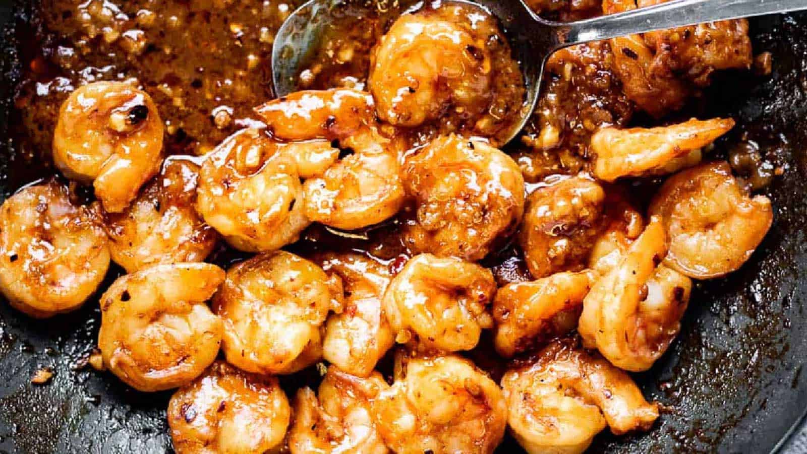 A close-up of cooked shrimp in a pan with a thick, dark brown sauce. The shrimp are evenly coated, and a spoon is seen lifting some sauce from the pan. This savory seafood preparation looks expertly made, almost as if it were auto-drafted by a culinary genius.