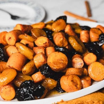 A close-up image of a white plate filled with glazed carrots, small potatoes, and prunes. A serving spoon is partially visible in the dish. In the background, there are two empty plates with silver forks on a marble surface.