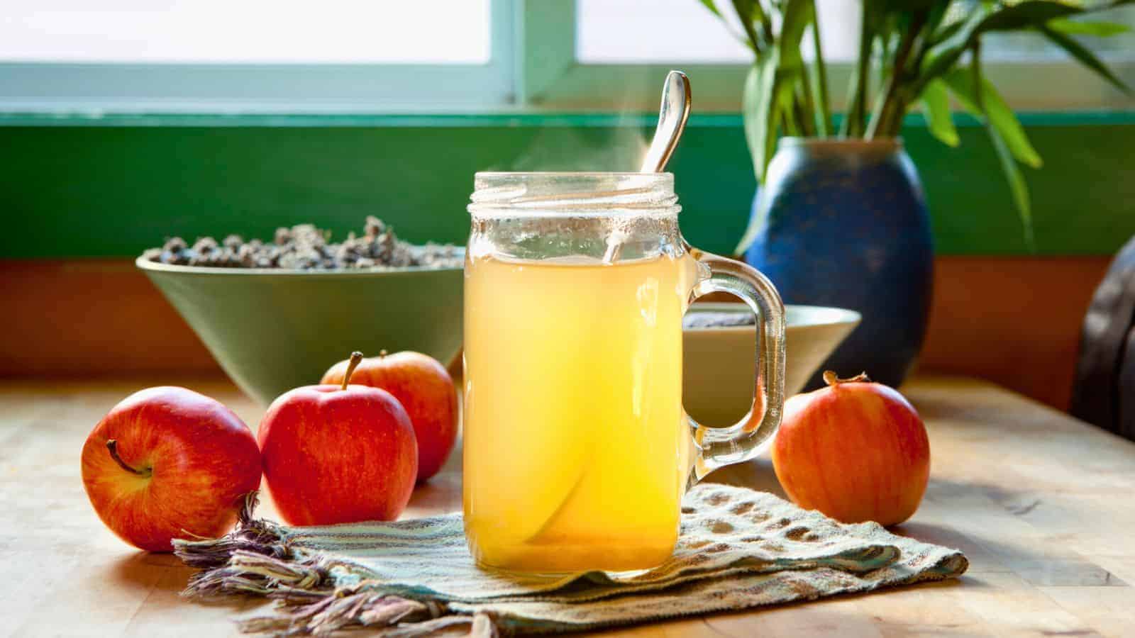A mug of hot apple cider with a spoon inside sits on a cloth on a wooden table, surrounded by three red apples. In the background, there is a window with green framing, a bowl containing dried lavender, and a blue vase with plants.