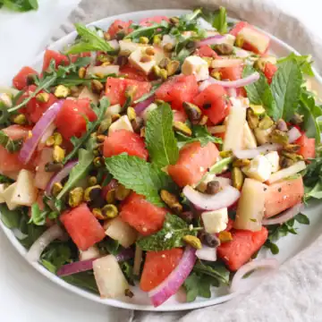 A plate of watermelon salad with arugula, sliced red onions, feta cheese cubes, chopped apples, pistachios, and fresh mint leaves. A wooden spoon and beige cloth are placed beside the plate on a white surface—a true summer blockbuster finale in the realm of watermelon recipes.