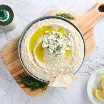 A bowl of Greek-inspired white bean hummus topped with crumbled feta cheese, olive oil, and dill sits on a wooden board. Two pieces of pita bread rest on the edge of the bowl. Nearby, a bottle and plate of lemon wedges are set on a marble surface.