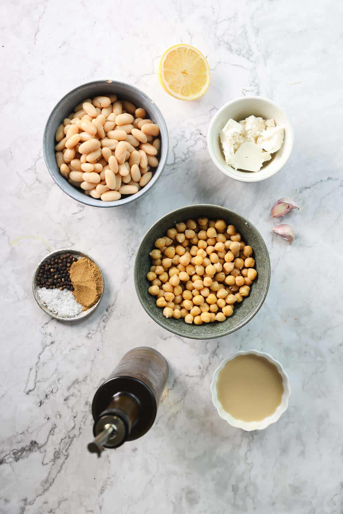 An overhead view of various ingredients on a marble surface, including a bowl of Greek-inspired white bean hummus, a bowl of chickpeas, a bowl with light-colored paste, feta crumbles in a small bowl with spices, a bottle of oil, three garlic cloves, a lemon half, and another bowl with white substance.