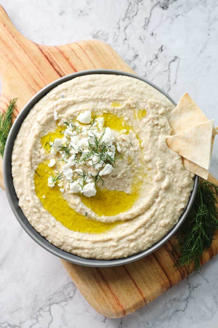 A bowl of Greek-inspired, creamy white bean hummus topped with crumbled feta cheese, olive oil, and dill sits on a wooden cutting board. Two pita bread pieces rest on the side of the bowl. The cutting board is placed on a marble surface.