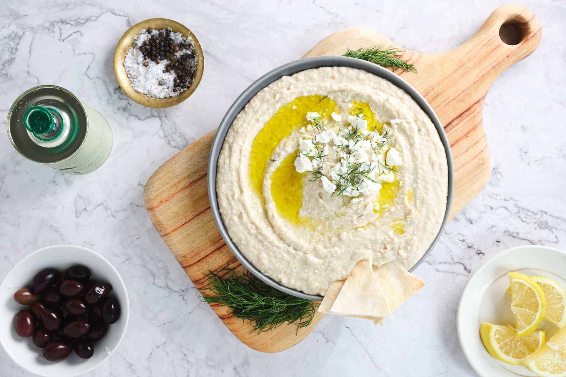 A bowl of Greek-inspired white bean hummus garnished with olive oil, chopped herbs, and feta cheese is placed on a wooden cutting board. Next to it are a bowl of black olives, a plate of lemon slices, a small bowl of salt and pepper, and a green bottle on a marble surface.