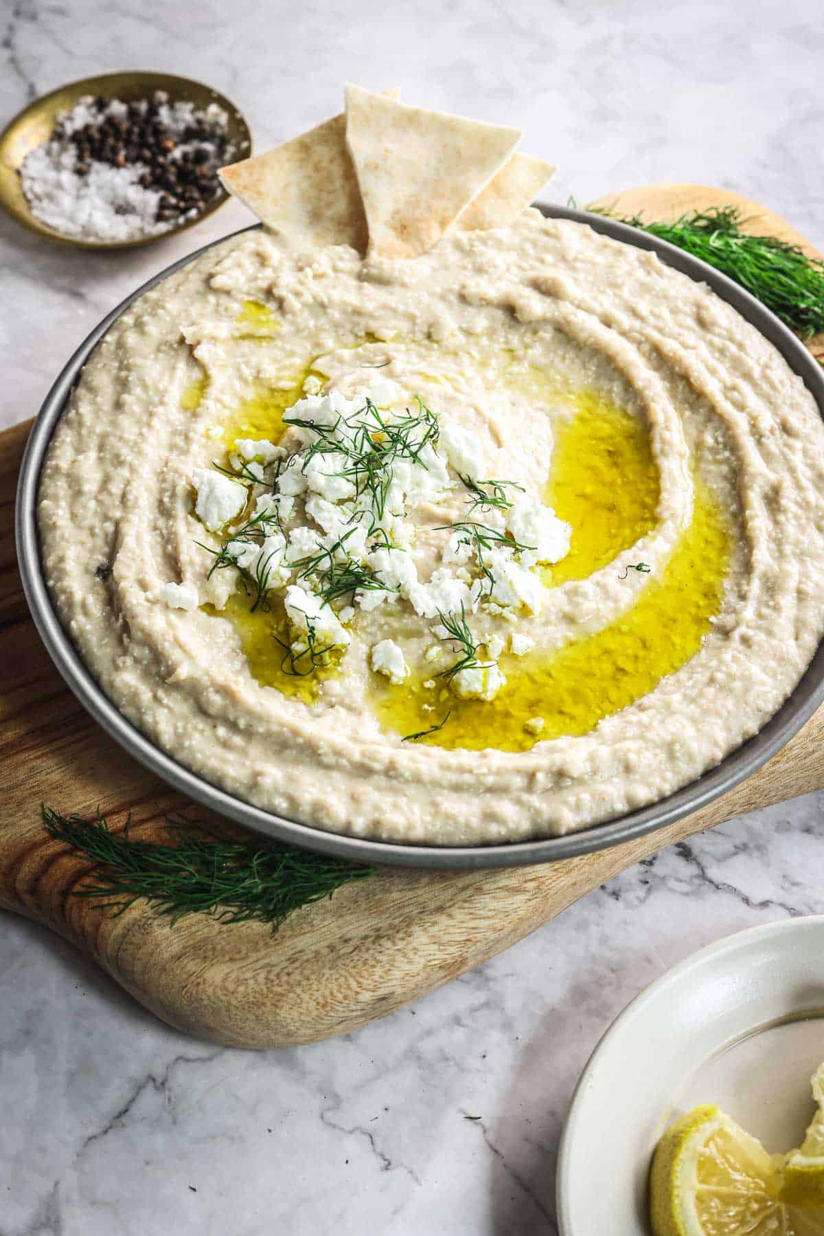 A bowl of creamy, Greek-inspired hummus topped with crumbled feta cheese, olive oil, and fresh dill sits on a wooden board. Two pita chips are dipped into the hummus. A lemon wedge and a small bowl with salt and pepper are in the background on a marble surface.