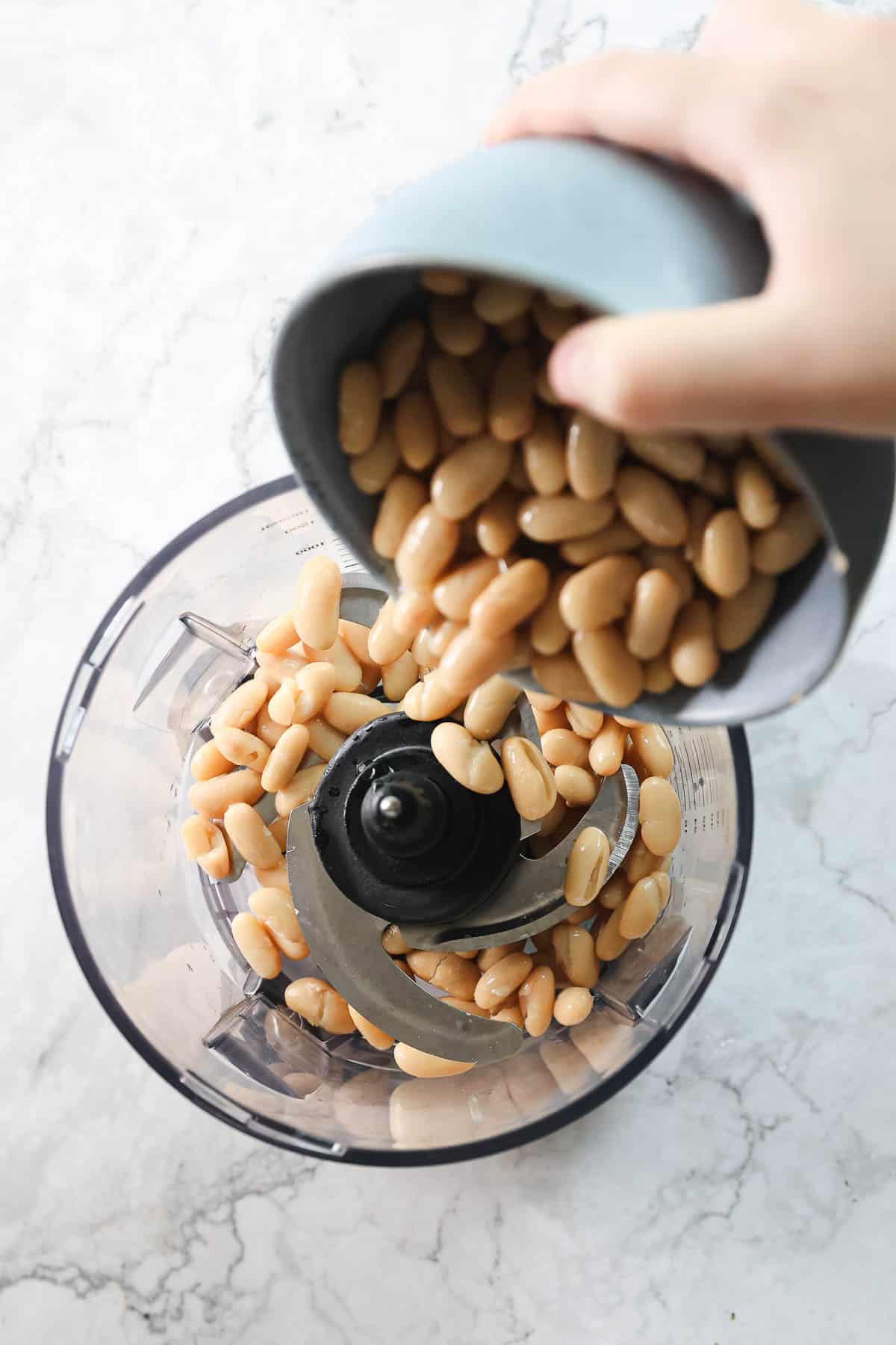 A person pours soaked white beans from a light blue bowl into a food processor with a metal blade attachment, ready to create creamy white bean hummus. The background shows a white marble surface.