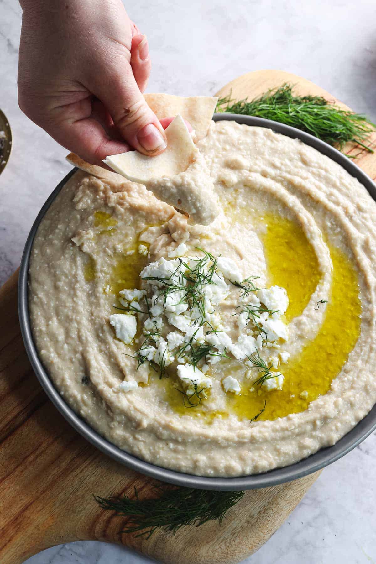 A hand dipping pita bread into a bowl of creamy white bean hummus topped with crumbled feta cheese, fresh dill, and a drizzle of olive oil. The bowl is placed on a wooden cutting board with dill sprigs around it.