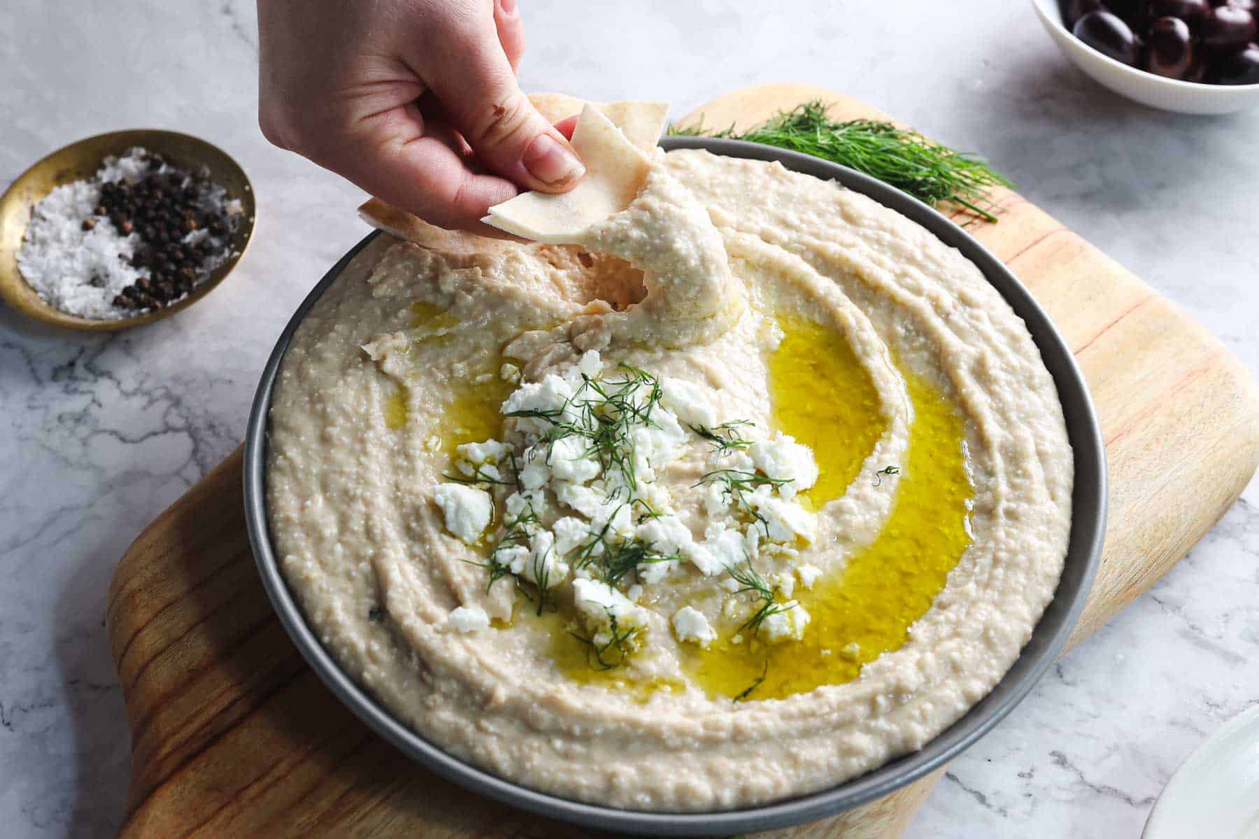 A hand dips a piece of pita bread into a bowl of Greek-inspired hummus topped with olive oil, crumbled feta cheese, and fresh herbs. The bowl is on a wooden cutting board, with a small dish of olives in the background and another small bowl containing salt and pepper.