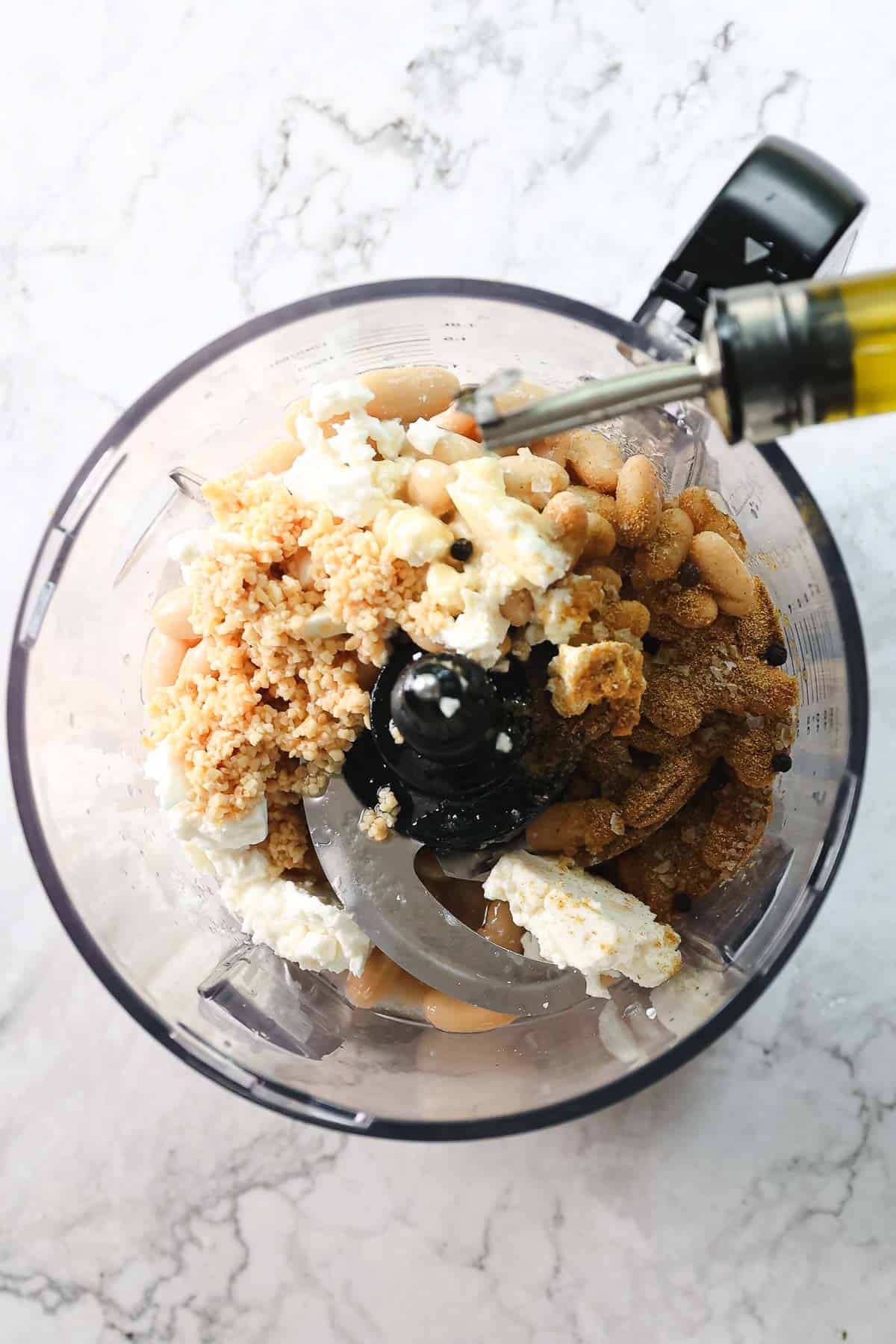 A food processor is shown from above, containing white beans, crumbled feta cheese, and brown sugar among other ingredients essential for a Greek-inspired white bean hummus. Olive oil is being poured into the mix against the backdrop of a pristine white marble countertop.