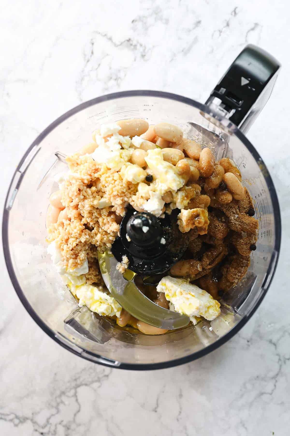 A top-down view of a food processor containing various ingredients, including white beans, crumbled feta cheese, and a brown crumbly mixture, set on a marble countertop. Some liquid is visible at the bottom of the processor, hinting at a Greek-inspired creamy white bean hummus.