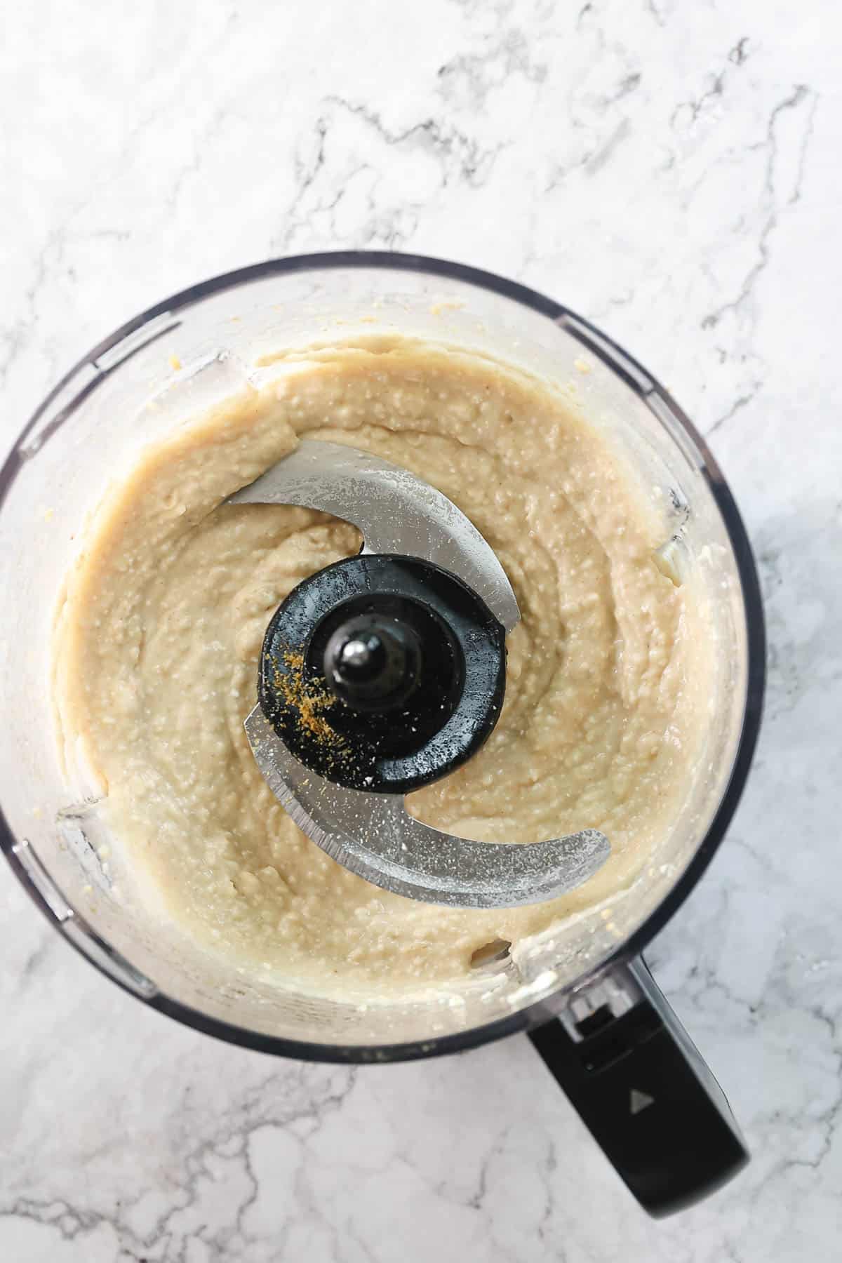 A top view of a food processor containing a creamy, beige mixture of Greek-inspired white bean hummus. The blending blade is visible in the center, and the mixture appears to be evenly blended. The processor is placed on a white and gray marbled countertop.