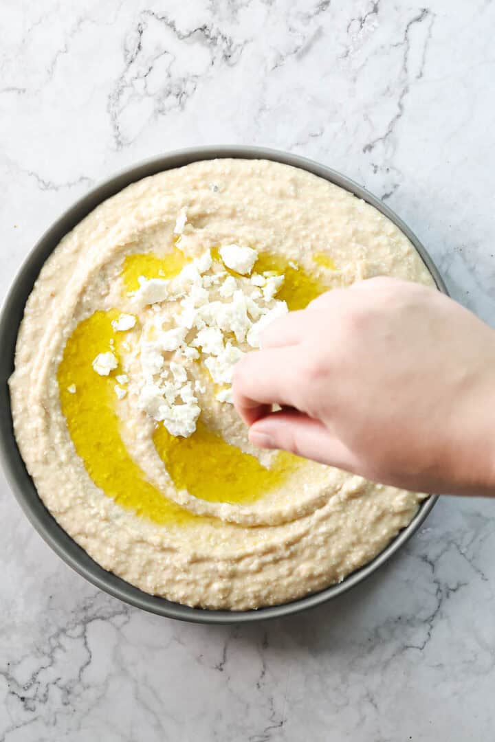 A bowl of Greek-inspired white bean hummus with a swirl of olive oil on top is being garnished with crumbled feta by a hand. The bowl is placed on a marble countertop.