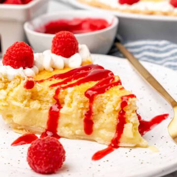 A slice of fruit-loaded lemon pie topped with whipped cream and raspberry sauce sits on a white plate. Fresh raspberries crown the slice, with extras scattered around. A gold fork rests beside it, while a bowl brimming with raspberries and a whole pie linger invitingly in the background.