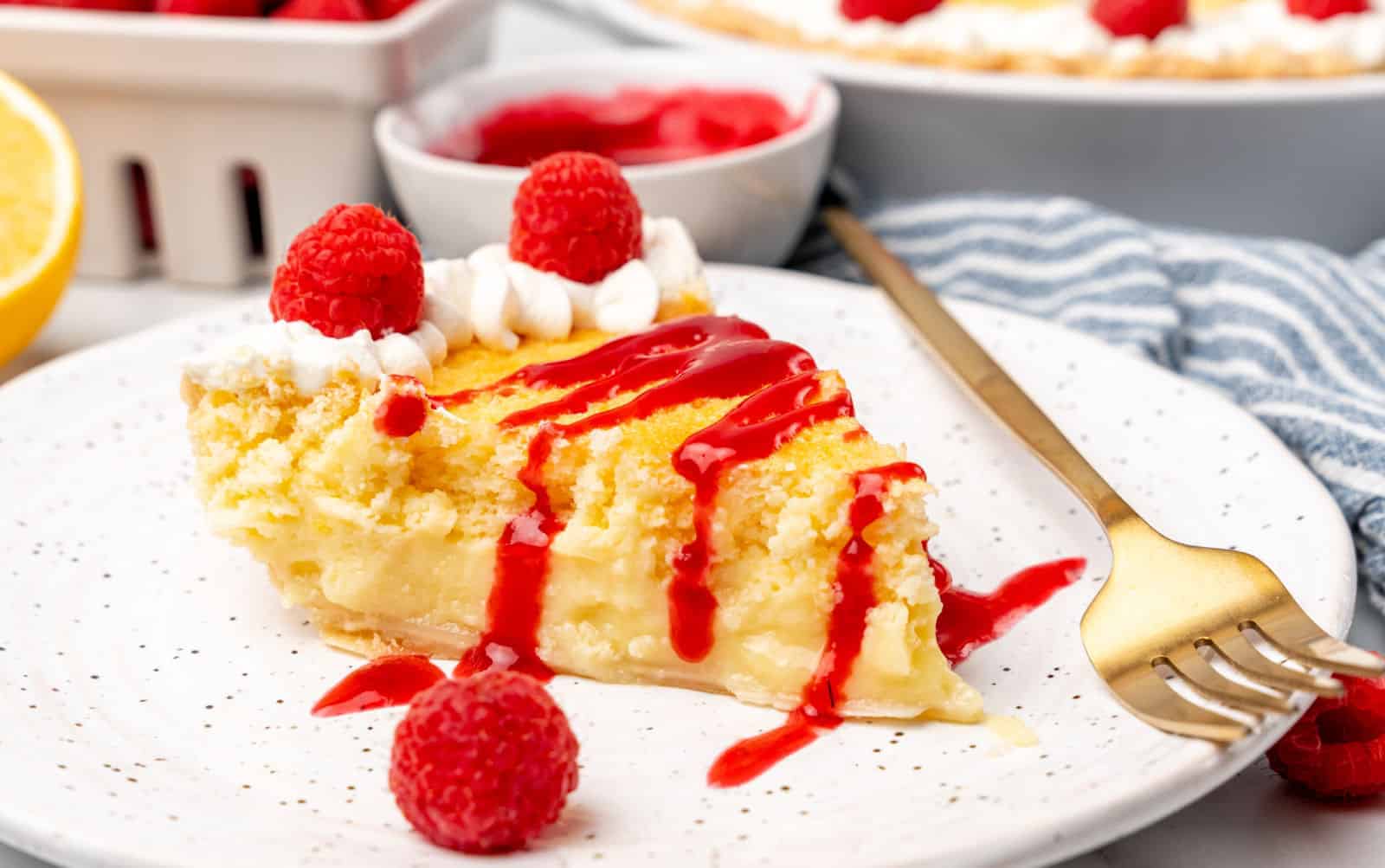 A slice of fruit-loaded lemon pie topped with whipped cream and raspberry sauce sits on a white plate. Fresh raspberries crown the slice, with extras scattered around. A gold fork rests beside it, while a bowl brimming with raspberries and a whole pie linger invitingly in the background.