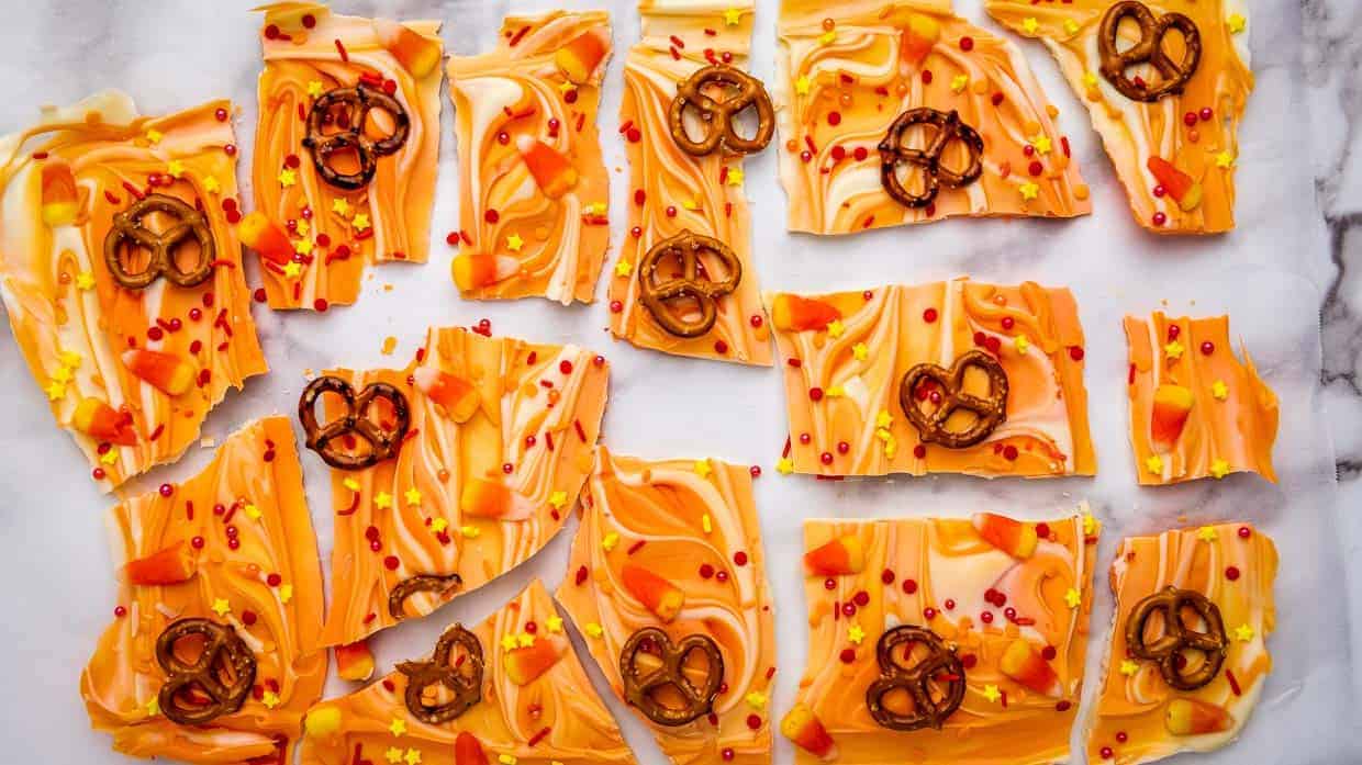 Rectangular pieces of orange and white swirled candy bark are topped with small pretzels, candy corn, and orange and yellow sprinkles. The pieces are arranged haphazardly on a white marble surface, making it one of the easiest Halloween recipes to impress your guests.
