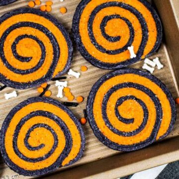 Cookies on a baking tray with orange and black swirled patterns. The tray is sprinkled with small orange, black, and white bone-shaped decorations. Perfect for your collection of easy Halloween recipes, the background is a white marble surface.