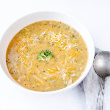 A bowl of creamy broccoli cheese soup garnished with shredded cheese and a small broccoli floret. A metal soup spoon rests on a light-colored napkin beside the bowl, ready for an auto draft of flavor. The background is white.