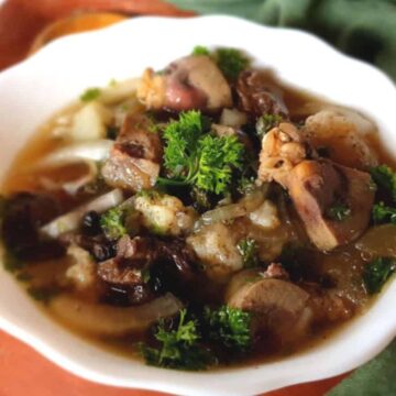 A white bowl filled with a hearty meat and vegetable stew, garnished with fresh parsley, sits on an orange surface. In the background, a green cloth and wooden cutlery add charm to the cozy scene—a perfect Auto Draft for comfort food inspiration.