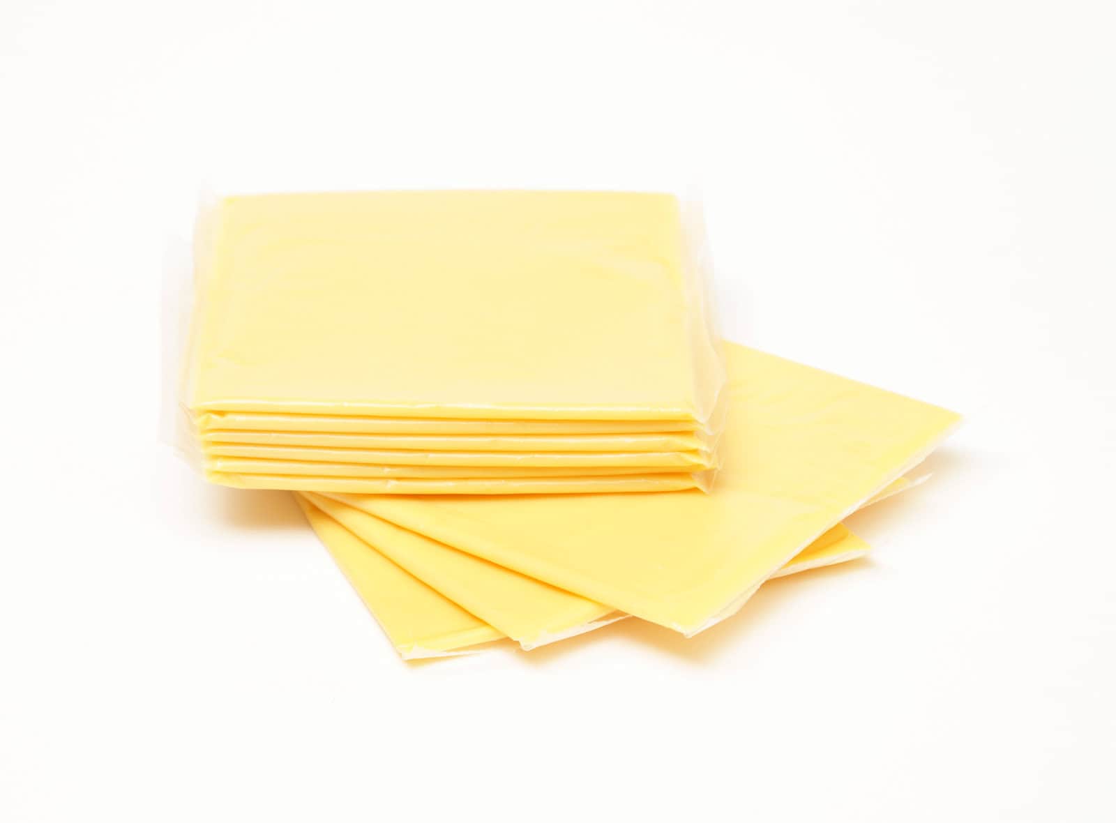 A stack of individually wrapped yellow cheese slices, a quintessential grocery item with a lot of chemicals, is arranged on a white surface. Four slices are fanned out in front of the stack, each in clear plastic wrapping.