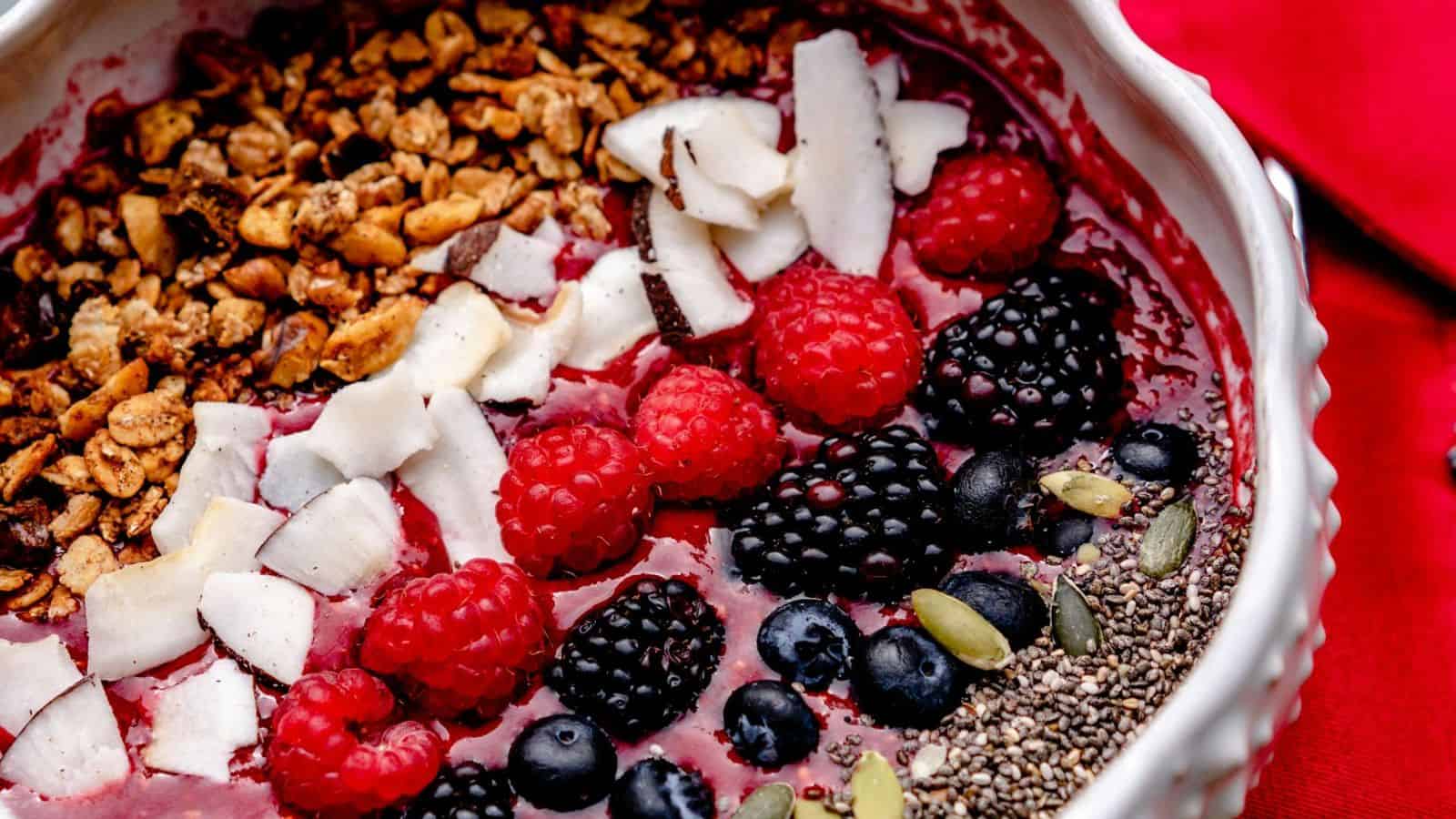 A bowl filled with a smoothie base topped with granola, shredded coconut, raspberries, blackberries, blueberries, chia seeds, and pumpkin seeds. The vibrant ingredients are arranged neatly and the bowl is set against a red background.