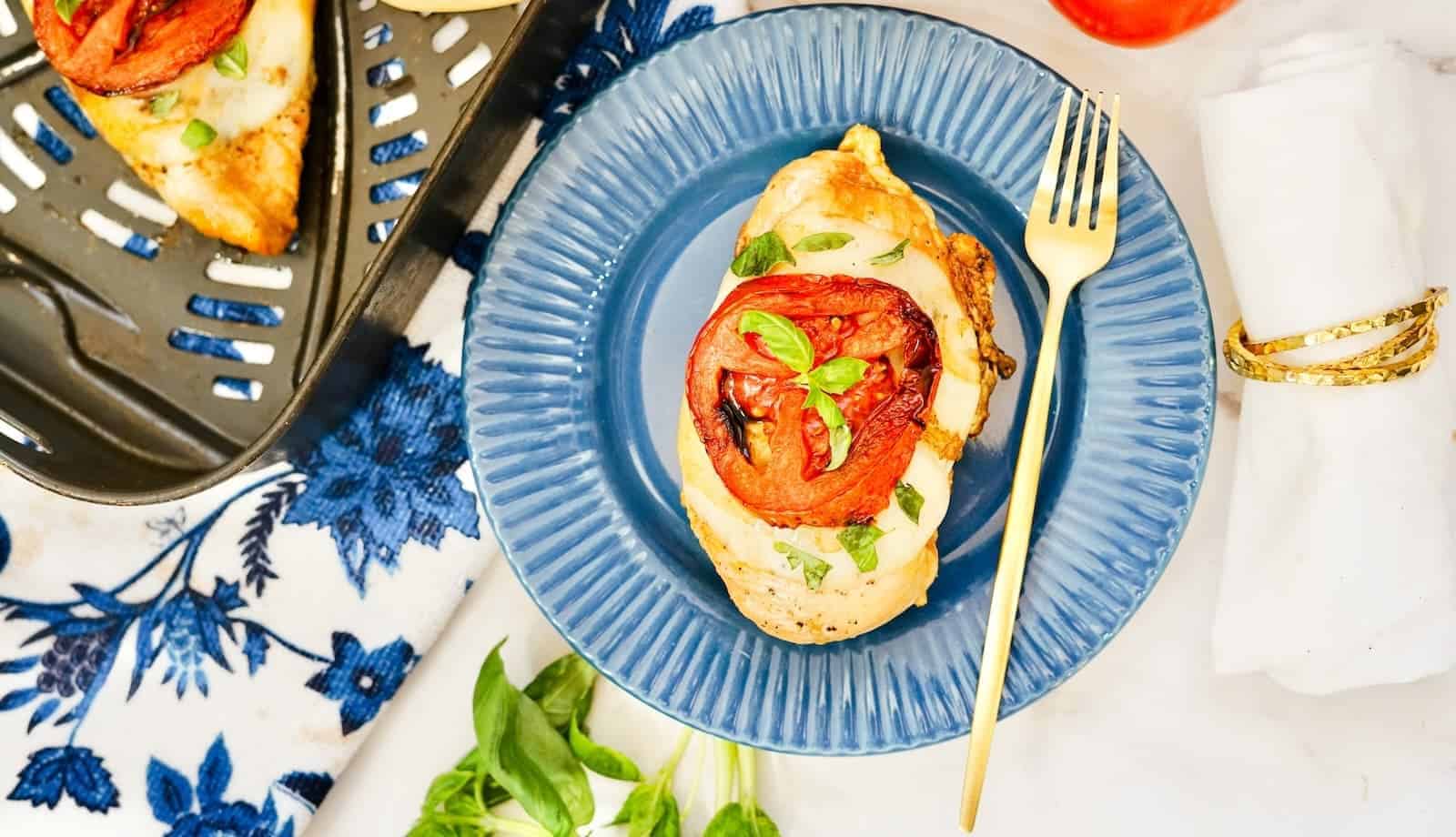 A cooked chicken breast topped with a slice of tomato and fresh basil sits on a blue plate, like an auto draft of culinary perfection. A gold fork rests beside it. Nearby, a white napkin with gold rings, a patterned cloth, and a grill tray with more chicken complete the scene.