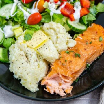 A plate of food featuring grilled salmon, mashed potatoes with butter, and a fresh salad with lettuce, cucumber, cherry tomatoes, and red peppers, drizzled with dressing. The dish is served on a dark plate.