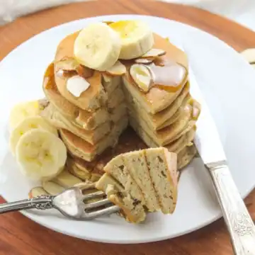A stack of pancakes topped with banana slices and syrup sits on a white plate. A slice has been cut out and is on a fork beside the stack. Almond slices are scattered on and around the pancakes, creating an enticing scene for pancake recipes enthusiasts. The plate rests on a wooden surface.