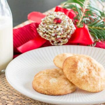 A white plate with three cookies is placed on a woven mat. A glass bottle of milk with a straw stands to the left of the plate. In the background, there is a red napkin, a gold and red ornament, and pine branches, suggesting a festive holiday setting ideal for exploring grandma recipes or iconic desserts.
