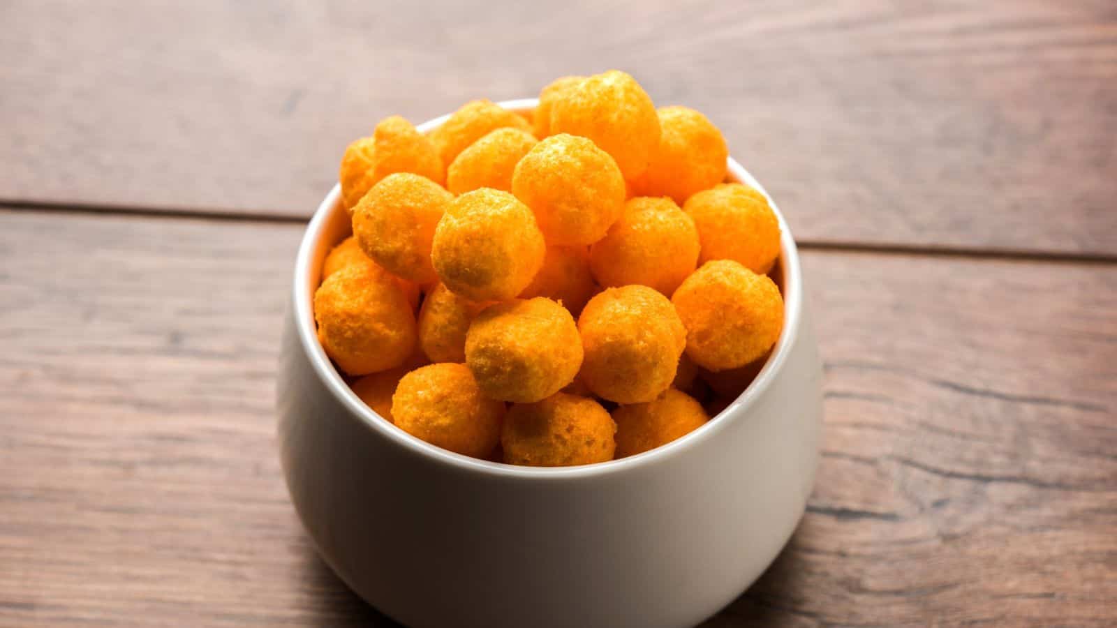 A white bowl filled with orange cheese puffs is placed on a wooden surface.