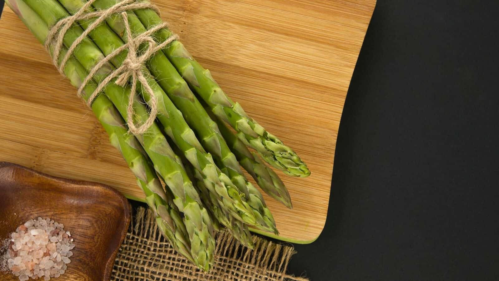 A bundle of fresh green asparagus tied with twine lies on a wooden cutting board. The board is placed on a black background, and a small wooden dish with pink salt is visible in the corner.