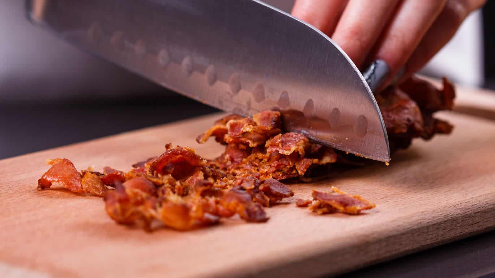 A hand holding a knife slices crispy bacon on a wooden cutting board. The bacon is in small pieces, and the surface is clean. The focus is on the knife and the bacon.